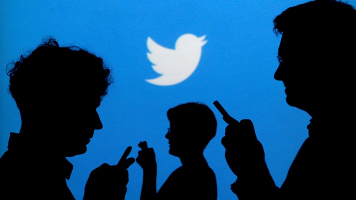 People in silhouettes look at their phones in front of a Twitter logo.