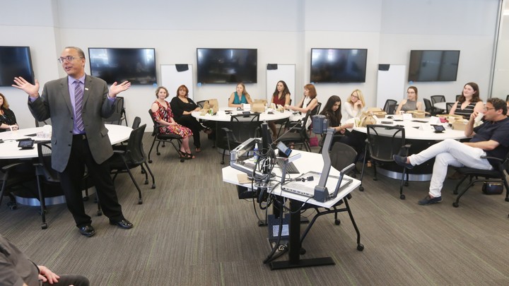 Students and professors sit around tables as one professor speaks.