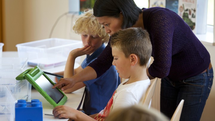 A teacher stands next to two students and looks at something on an iPad with them.