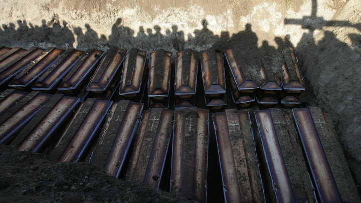 Shadows of people burying dozens of coffins in a mass grave.