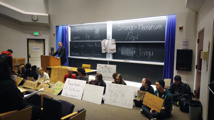 A professor stands at a lecture podium, while protesters sit alongside him holding signs.