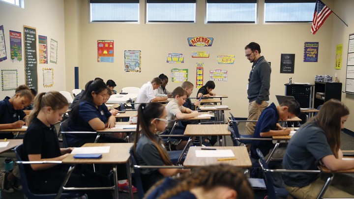 Students take a test in a classroom; a teacher stands at the front of the room.