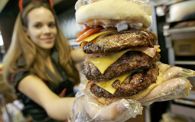 A restaurant worker in a red and black uniform holds a three-patty cheeseburger.