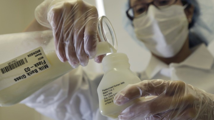 A person wearing gloves and a surgical mask pours liquid from a flask labeled "Milk Bank" into a jar