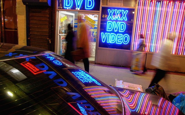 The neon lights of an adult-video store are reflected on a car.