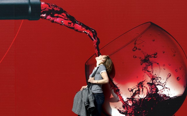 A woman stands with her mouth open in front of a large illustration of red wine being poured into a glass.