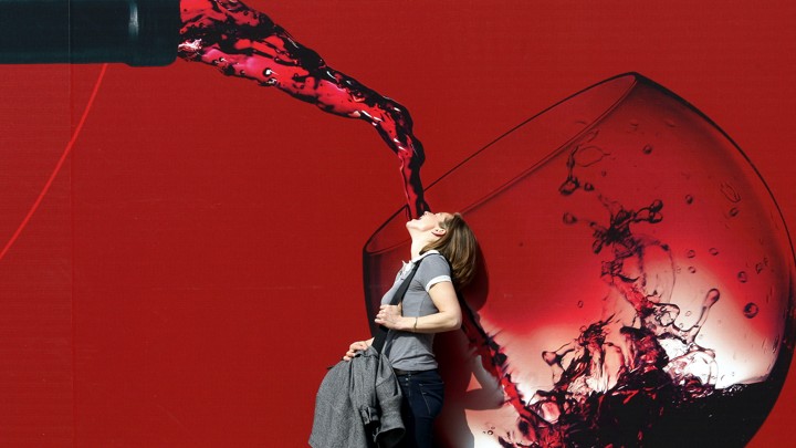 A woman stands with her mouth open in front of a large illustration of red wine being poured into a glass.