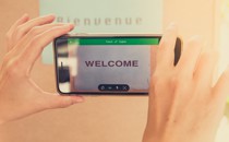 Hands hold a smartphone in front of a sign saying "Bienvenue" and the smartphone reads "Welcome"