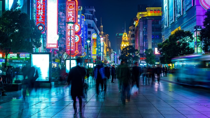 Blurred individuals walk on a street lit by colorful signs.