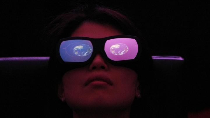 A person wears large glasses with one red and one blue lens and an image of the Earth reflected in each.
