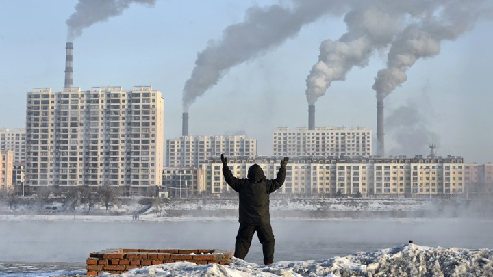 A photograph of a person facing away from the camera, standing with their arms raised, in front of factories with smokestacks emitting smoke