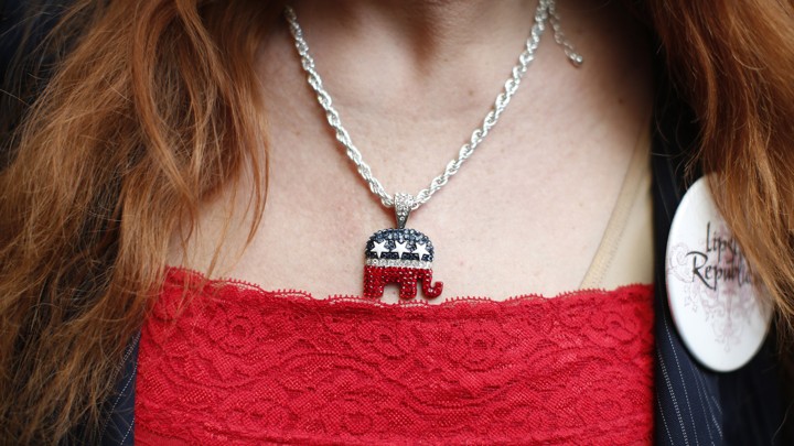 A close-up photograph of a woman wearing a red shirt and a sparkly necklace with a Republican elephant pendant