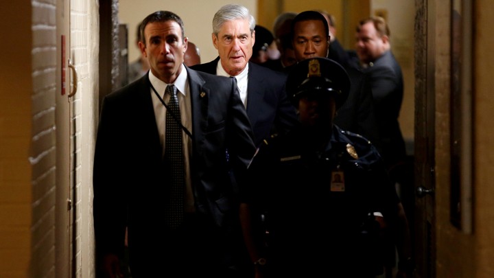 Special Counsel Robert Mueller and other officials walk down a hallway accompanied by a police officer.