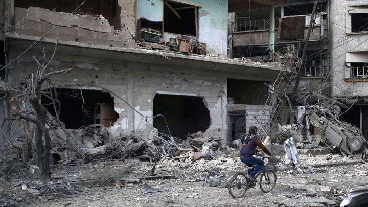 A boy rides his bicycle past the rubble of bombed-out buildings.