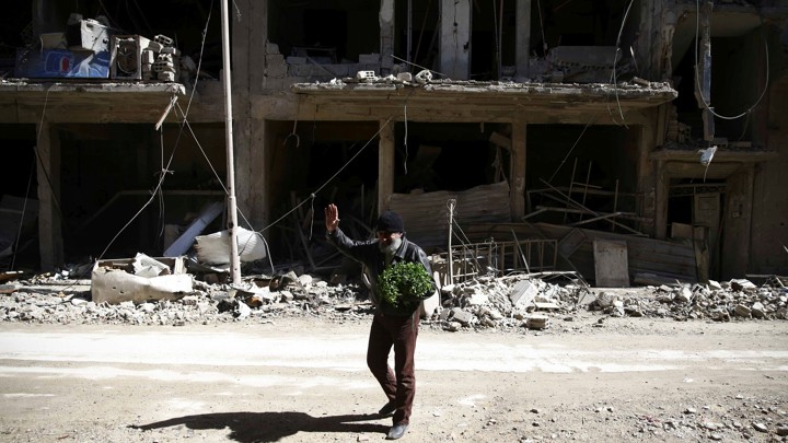A bearded man waves outside the shell of a bombed-out building.