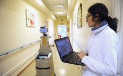A human caregiver uses a laptop to operate a robot caregiver in the hallway of a nursing residence.