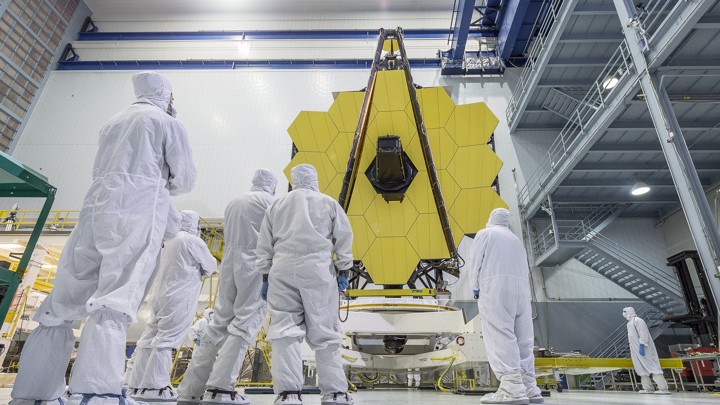 NASA employees, dressed in white uniforms, look up at the gold-plated mirrors of the James Webb Space Telescope.