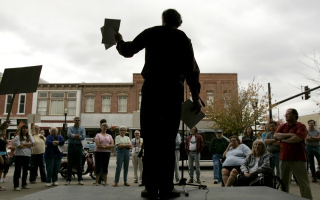 Al Norman, seen in silhouette, holds flyers and speaks to a crowd from a stage.