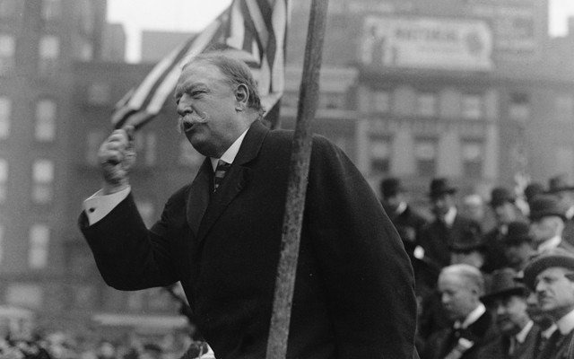 Former President William Howard Taft speaks at a campaign rally, with the American flag waving behind him.