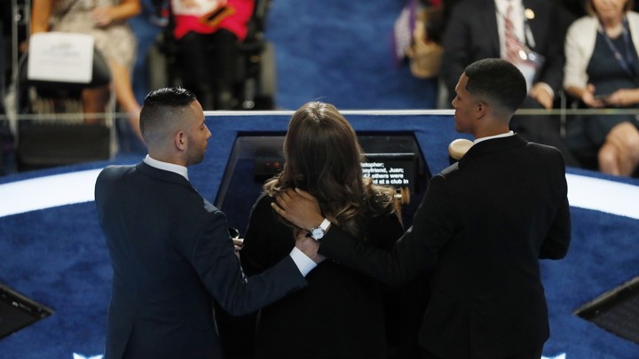 A view from behind of Christine Leinonen speaking at a podium, with Juan Arriagada and Brandon Wolf holding her shoulders