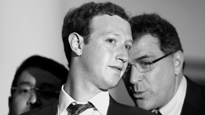 A black and white photograph of a bespectacled man whispering into Mark Zuckerberg's ear