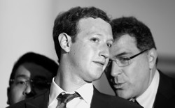 A black and white photograph of a bespectacled man whispering into Mark Zuckerberg's ear