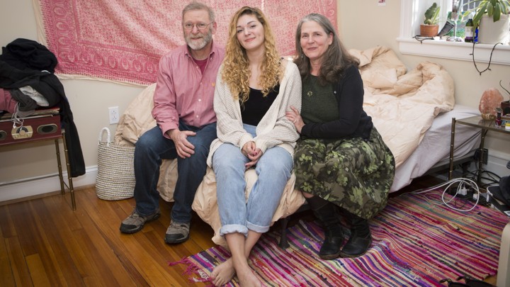Two parents and their young adult daughter sit on a bed in a college apartment.