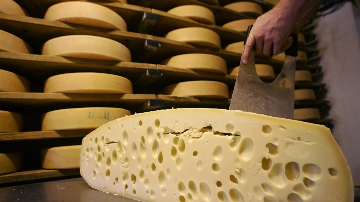 A hand slices cheese in front of shelves of more cheese.