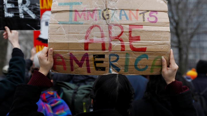 Hands holding a colorful sign that reads "Immigrants Are America"