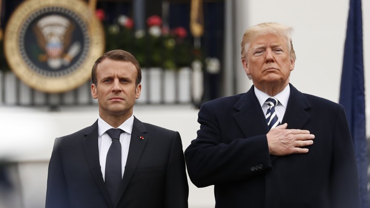 Macron standing with arms at sides next to Trump, who has a hand over his heart