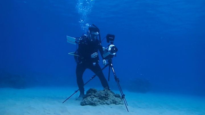 A diver wearing scuba equipment sets up a camera on a tripod underwater.