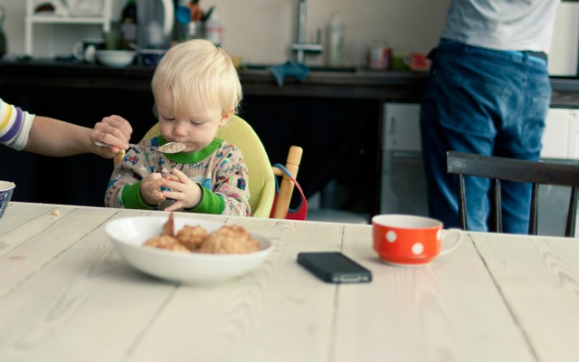 A mother spoon-feeds an infant while a man stands at a kitchen counter