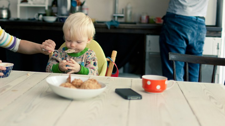 A mother spoon-feeds an infant while a man stands at a kitchen counter
