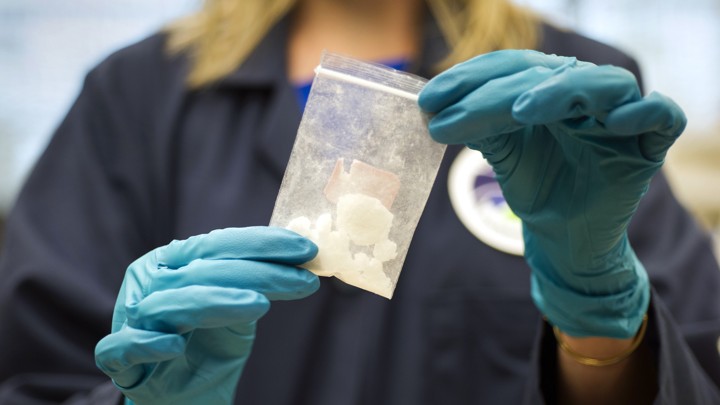 A person with gloved hands holds a small plastic bag of white powder.
