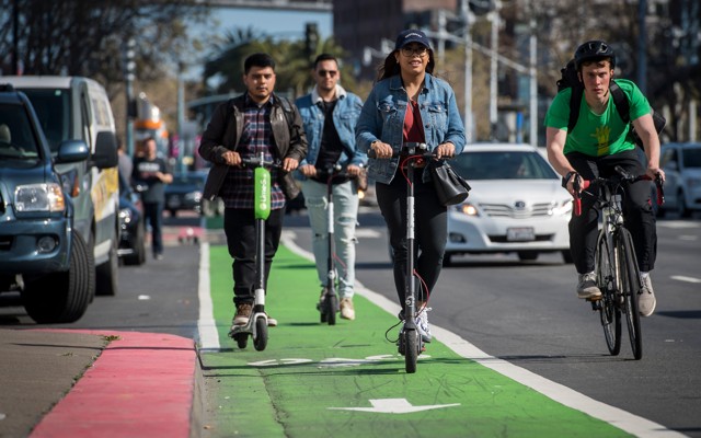 A group of people rides electric scooters from Lime and Bird in a bike lane while a cyclist passes them.