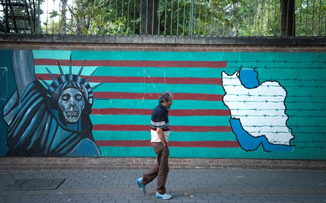 A man in front of an anti-U.S. mural featuring the Statue of Liberty, the American flag, and a map of Iran in barbed wire