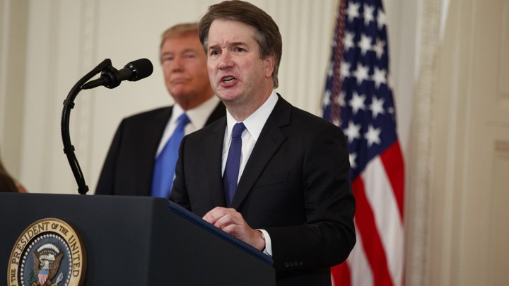 Brett Kavanaugh speaks at a podium with President Trump and an American flag in the background