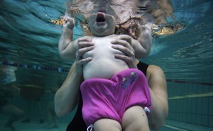 A baby is seen underwater from the chin down, held in the hands of an adult.