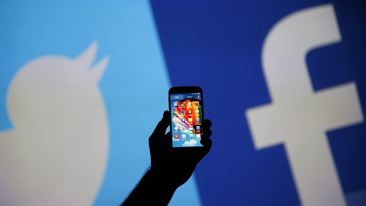A hand holds a smartphone up in front of Twitter's and Facebook's logo
