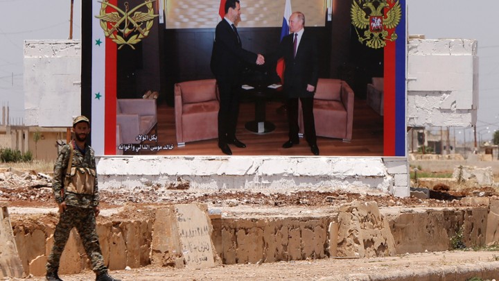 A soldier stands on a road in Syria in front of a billboard with a photo of Assad and Putin shaking hands.