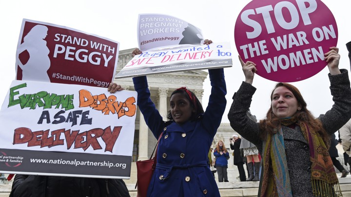 Three women holding signs stand in front of the Supreme Court, protesting a decision