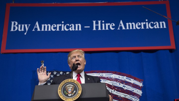 Donald Trump stands behind a podium with a banner behind him that says "Buy American - Hire American"