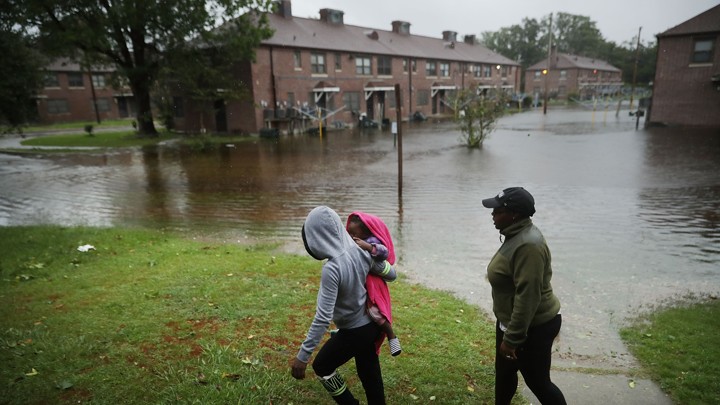 Two adults and a small child walk across a lawn with a flooded street in New Bern, North Carolina, behind them.