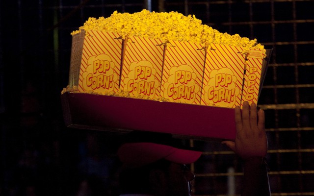 A vendor carries large bags of popcorn on a tray above his head