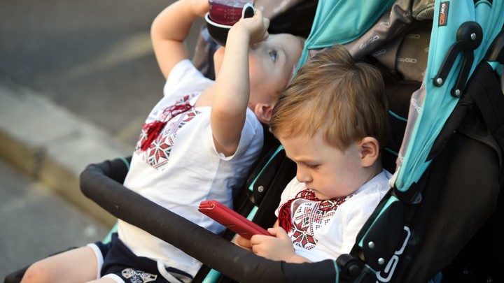 A young child plays with a phone while his brother drinks juice.