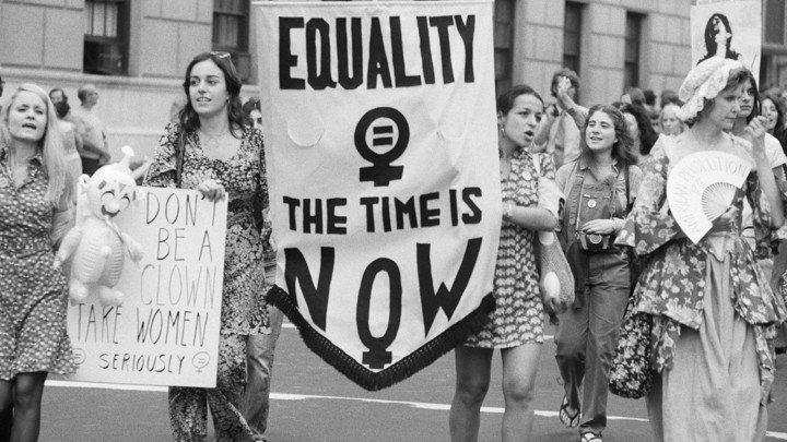 Women carry a sign reading "Equality: The Time Is Now" at a women's liberation parade.