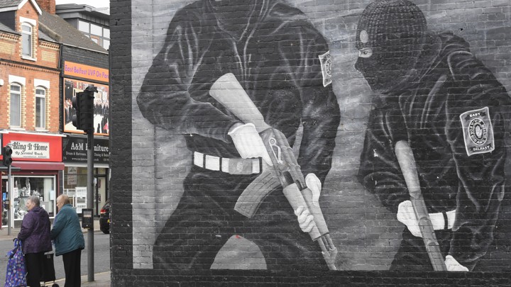 Pedestrians pass a political mural in Belfast, Northern Ireland, showing two men in balaclavas holding guns.