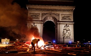 A protester wearing a yellow vest burns a barricade in front of a stone arch.