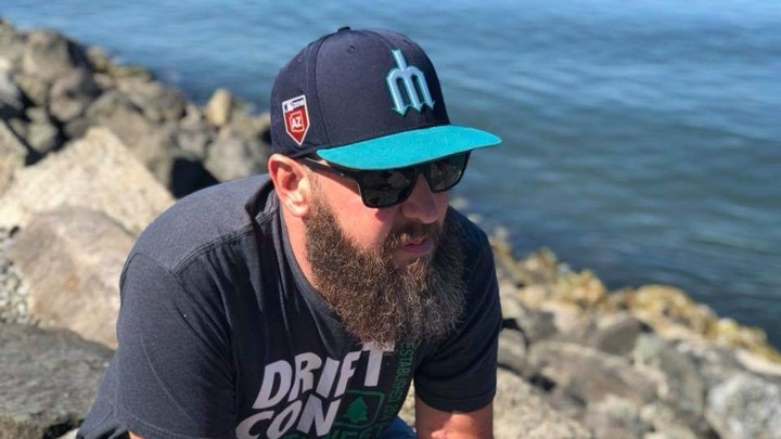 A bearded man in a baseball cap sits on some rocks by the water.