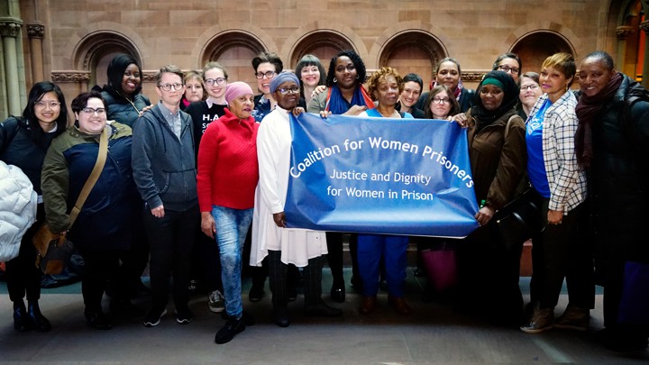 A group of women hold up a banner reading "Coalition for Women Prisoners: Justice and Dignity for Women in Prison"
